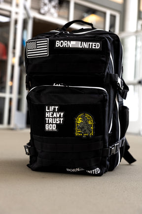 The Apex Premium Tac-Pac - 45L by Born United Private Label, a black tactical travel backpack with white trim, stands upright. Patches read “BORN UNITED,” “LIFT HEAVY TRUST GOD,” plus a U.S. flag and yellow graphic. Indoor setting blurred behind.