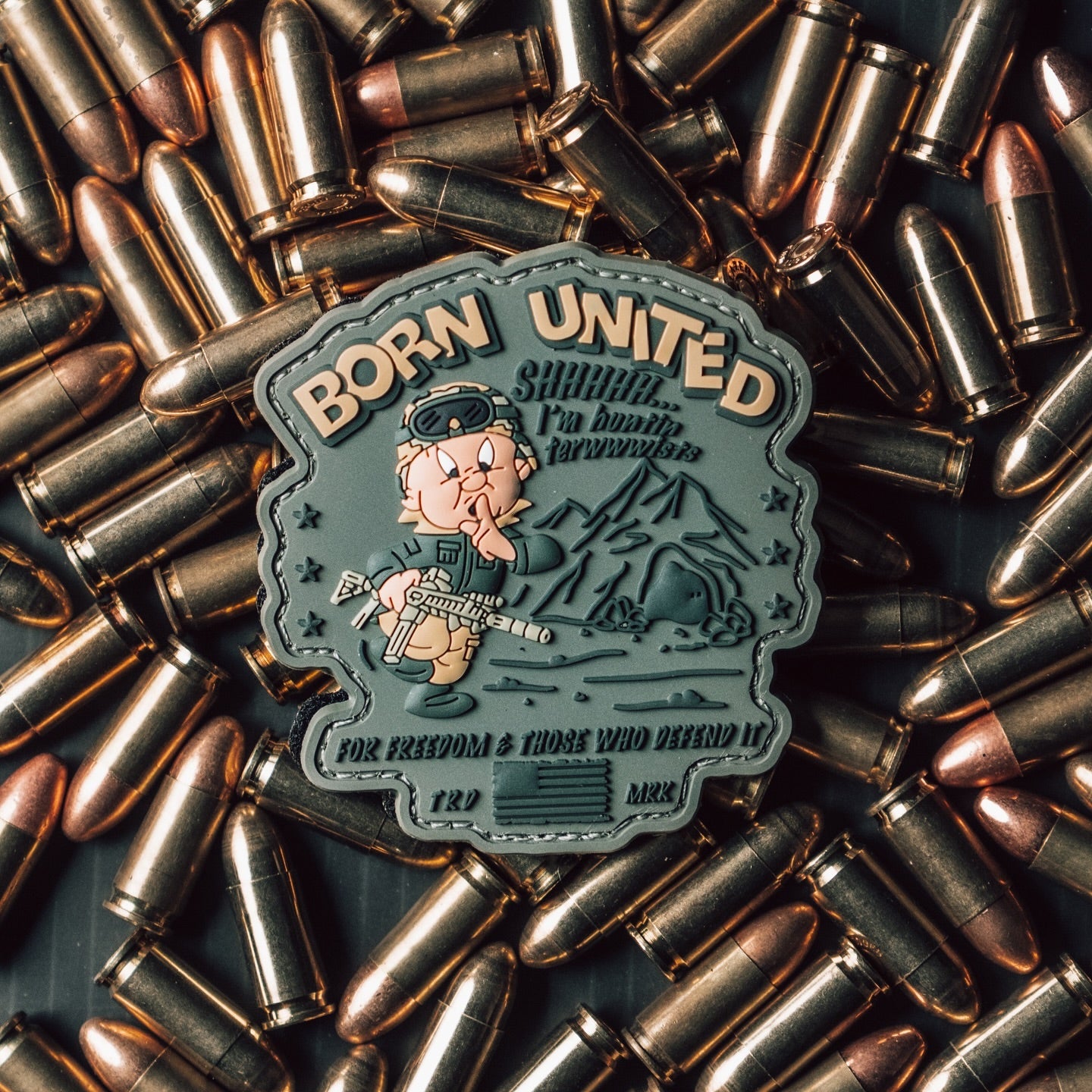 A patch with a cartoon character holding a rifle is surrounded by bullets. The patch reads BORN UNITED and features a camo-wearing figure against a mountain backdrop, with phrases and designs related to freedom and defense.