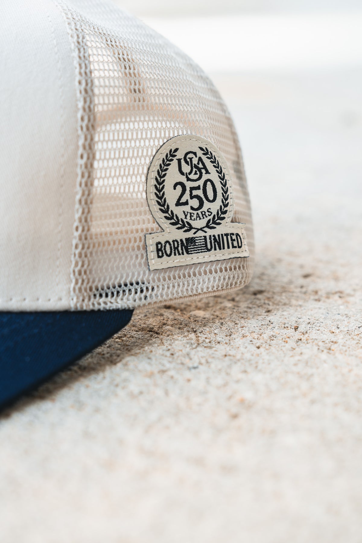 Close-up of the ’Merica Trucker Snapback by Born United Private Label, featuring a white mesh design and a patch that reads “USA 250 Years Born United,” displayed on a light textured surface. This breathable cap is ideal for those who value handmade hats offering both style and comfort.