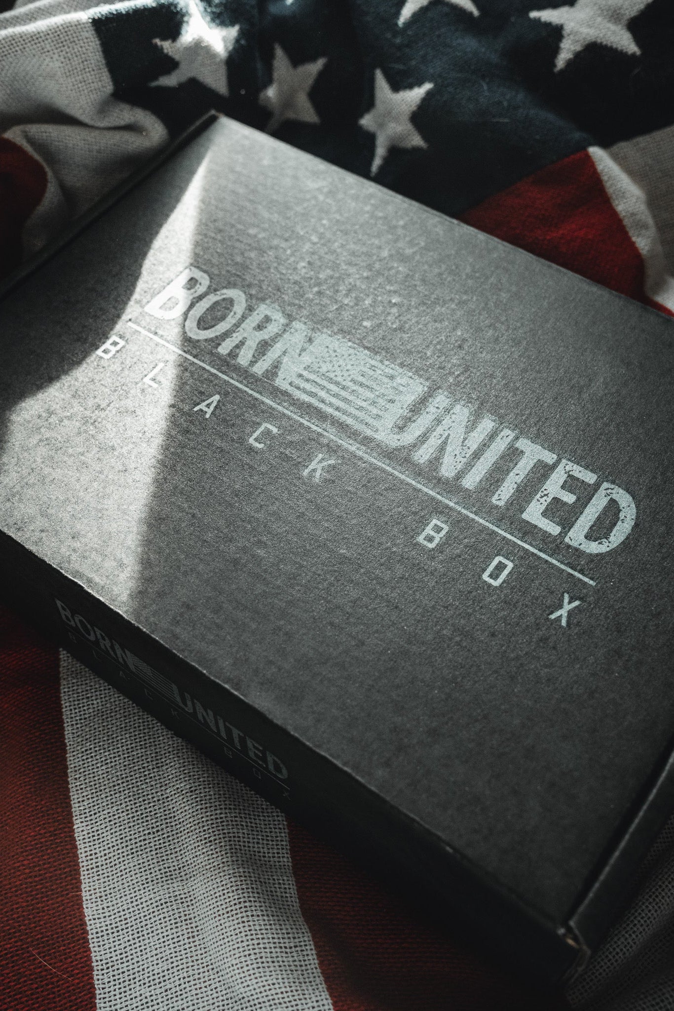 A black "Born United Black Box - Tactigator" by Born United Private Label rests on a partially visible American flag, hinting at exclusive or limited edition tees inside, with light and shadow highlighting the box and fabric.