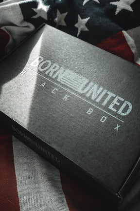 A black "Born United Black Box - Tactigator" by Born United Private Label rests on a partially visible American flag, hinting at exclusive or limited edition tees inside, with light and shadow highlighting the box and fabric.
