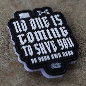 The "No One Is Coming To Save You - Patch" by Tango Charlie Apparel features bold white text, a small American flag, and crossed barbells on 3D PVC with Velcro backing for easy attachment, shown on a rough tan surface.