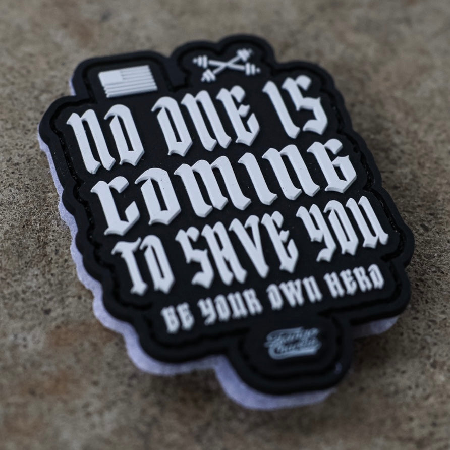 The "No One Is Coming To Save You - Patch" by Tango Charlie Apparel features bold white text, a small American flag, and crossed barbells on 3D PVC with Velcro backing for easy attachment, shown on a rough tan surface.