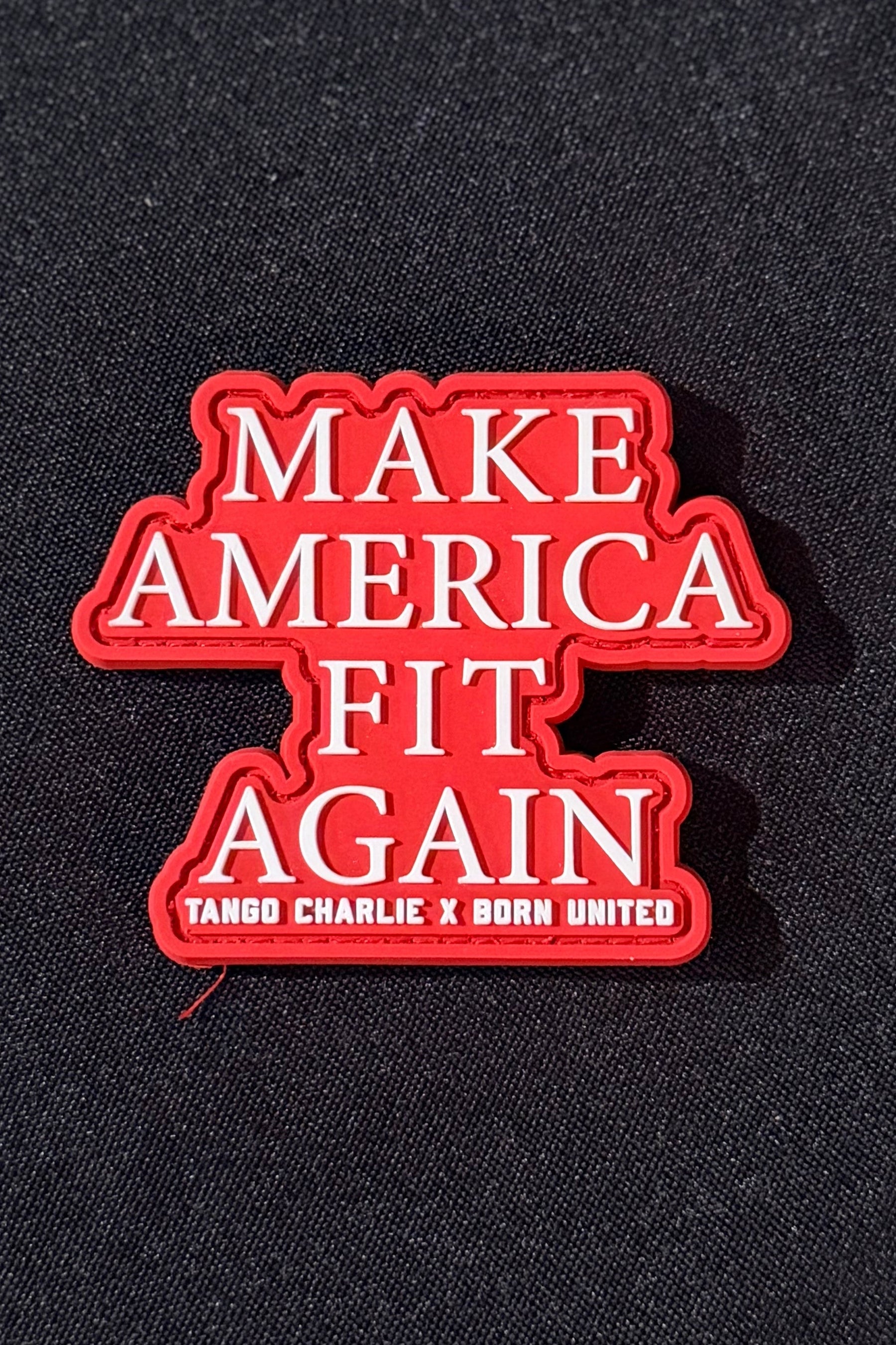 A red and white enamel pin with the text 'MAKE AMERICA FIT AGAIN' and 'TANGO CHARLIE X BORN UNITED' on a black textured fabric background.
