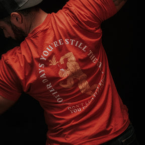 A person in a red St. Arnold The Stoic t-shirt by Tango Charlie Apparel, featuring a roaring tiger and the words “Other days you’re still the tiger. Don’t let anyone tell you different,” flexes with their back to the camera.