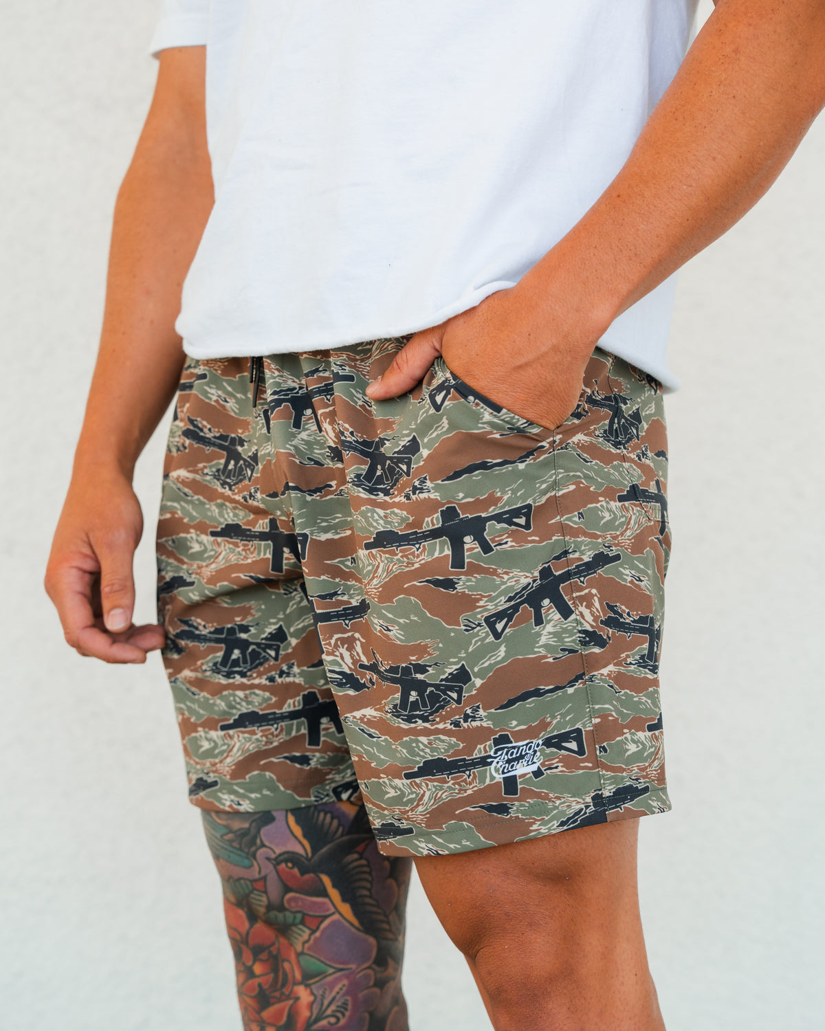 A person in a white T-shirt and Tango Charlie Apparel Men's Performance Training Shorts | 5.5" - Snipes & Stripes, featuring a rifle pattern, stands with one hand in their pocket. A colorful tattoo is visible on their left leg against a plain background.