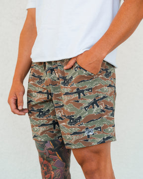 A person in a white T-shirt and Tango Charlie Apparel Men's Performance Training Shorts | 5.5" - Snipes & Stripes, featuring a rifle pattern, stands with one hand in their pocket. A colorful tattoo is visible on their left leg against a plain background.