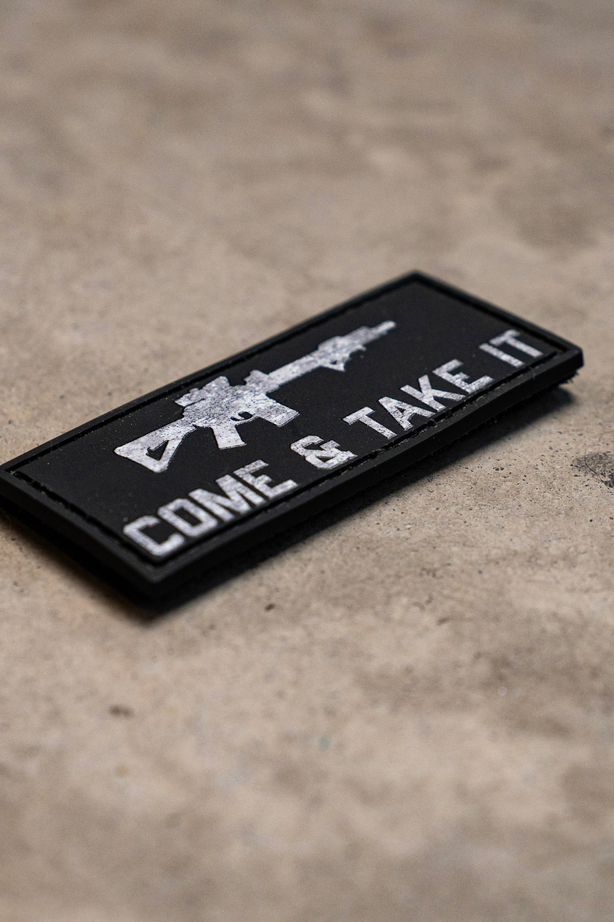 The Born United Private Label Come and Take It PVC Patch, featuring a white rifle and text on black, is displayed on a concrete surface.