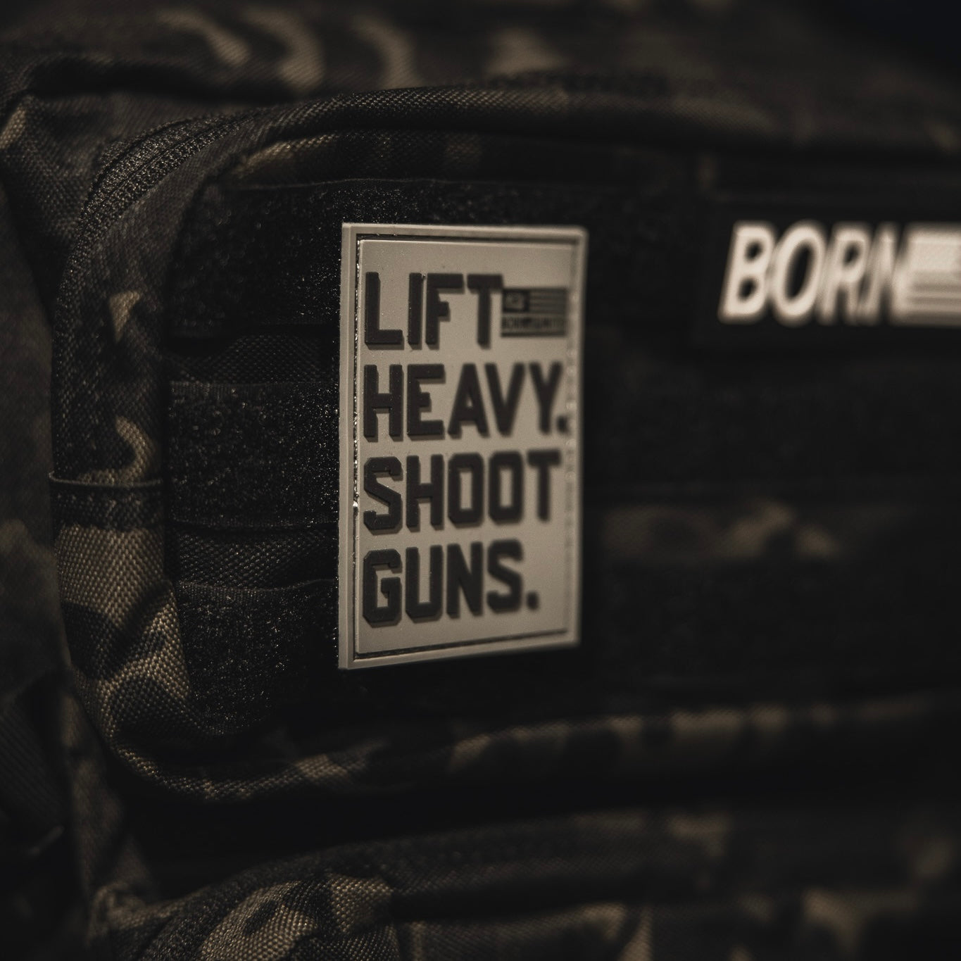A close-up of a dark camouflage backpack shows a bold black 3D PVC Velcro LHSG - Patch by Tango Charlie Apparel that reads, LIFT HEAVY. SHOOT GUNS. Another blurred patch with partial text is seen above it.