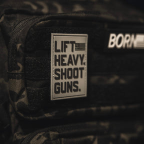 A close-up of a dark camouflage backpack shows a bold black 3D PVC Velcro LHSG - Patch by Tango Charlie Apparel that reads, LIFT HEAVY. SHOOT GUNS. Another blurred patch with partial text is seen above it.