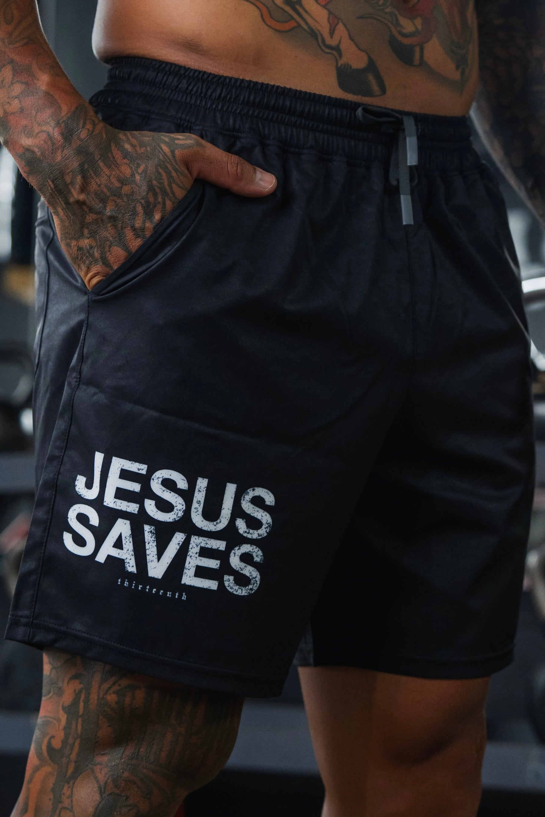 A tattooed individual wears 13th Men’s Standard Performance Shorts | 5.5"- Jesus Saves, featuring bold white lettering on the left leg. Their hand rests in their pocket, with a blurred background.