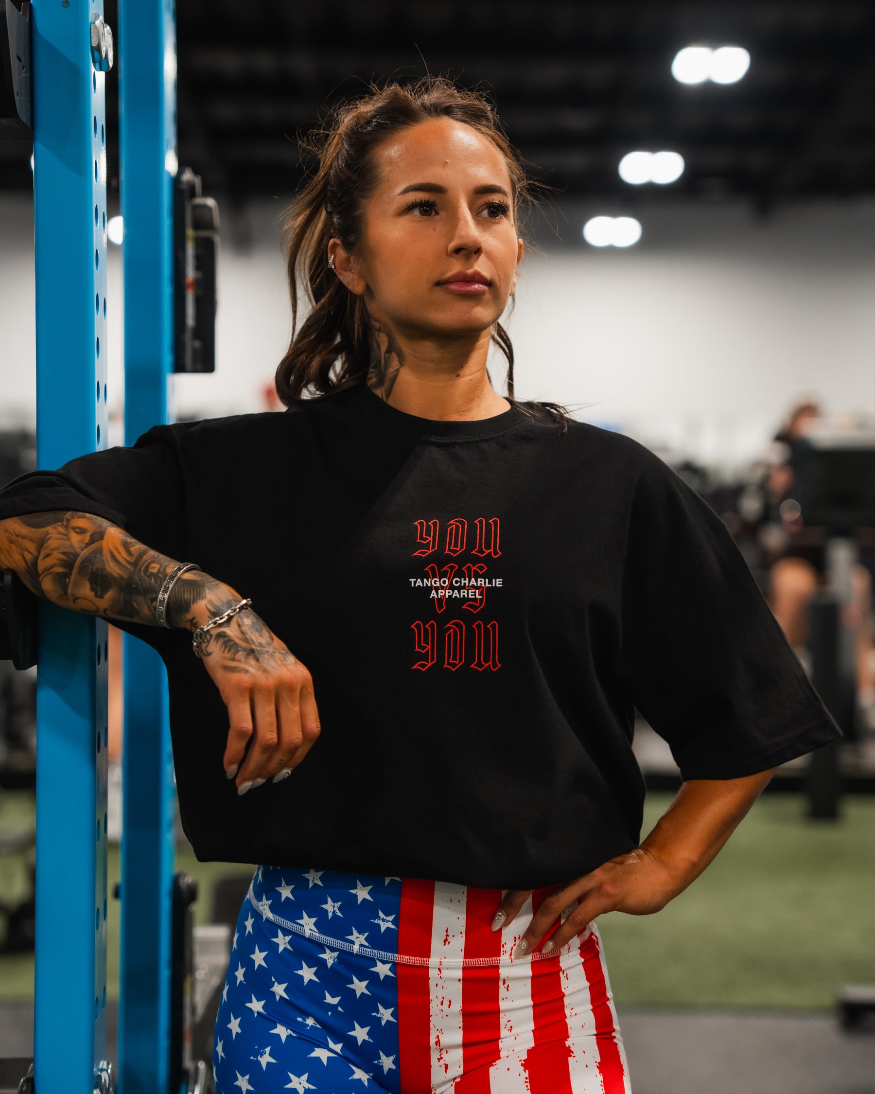A tattooed woman in a gym wears a black shirt with red print and Born United Private Label Women’s High Waisted Seamless Front Compression Training Shorts in Stars & Stripes. She leans on blue equipment; the background shows blurred gym gear and people.