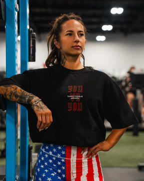 A tattooed woman in a gym wears a black shirt with red print and Born United Private Label Women’s High Waisted Seamless Front Compression Training Shorts in Stars & Stripes. She leans on blue equipment; the background shows blurred gym gear and people.