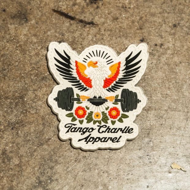 The "Old School, Never Gets Old - Patch" by Tango Charlie Apparel features an eagle with spread wings holding a barbell over red and yellow flowers, rays behind its head, and the brand name on a mottled grey background.