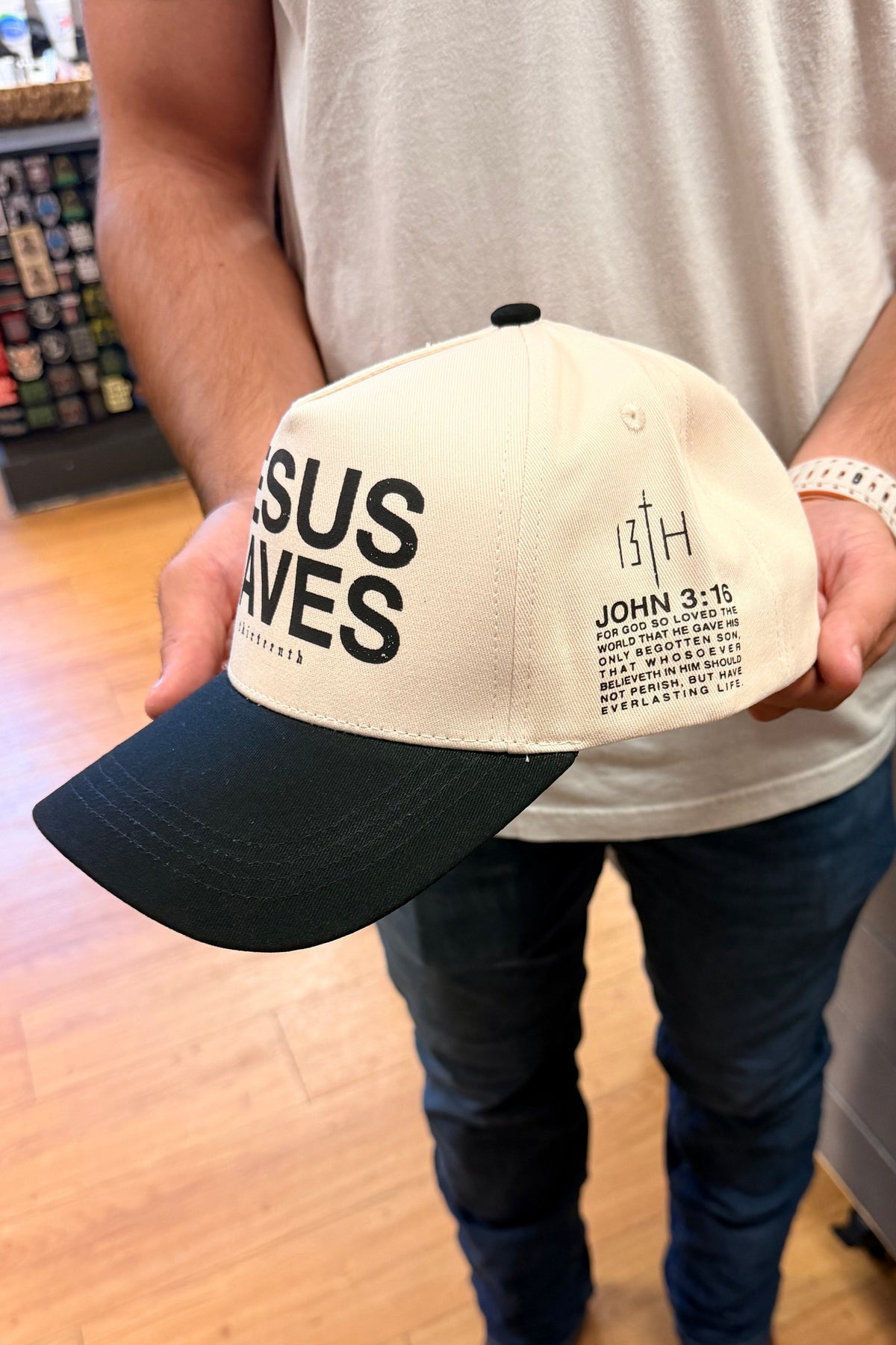 A man in a beige t-shirt and jeans holds up a baseball cap with a cream-colored crown and a black brim. The cap features the text 'JESUS SAVES' in black on the front and 'JOHN 3:16' with scripture text on the side.
