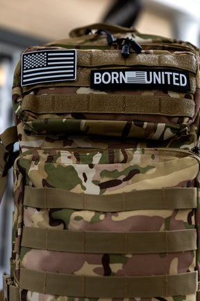 The Born United Private Label OCP Tac-Pac - 45L features a camouflage design, two front patches (black-and-white American flag and BORN UNITED), multiple straps, and a zippered pocket.