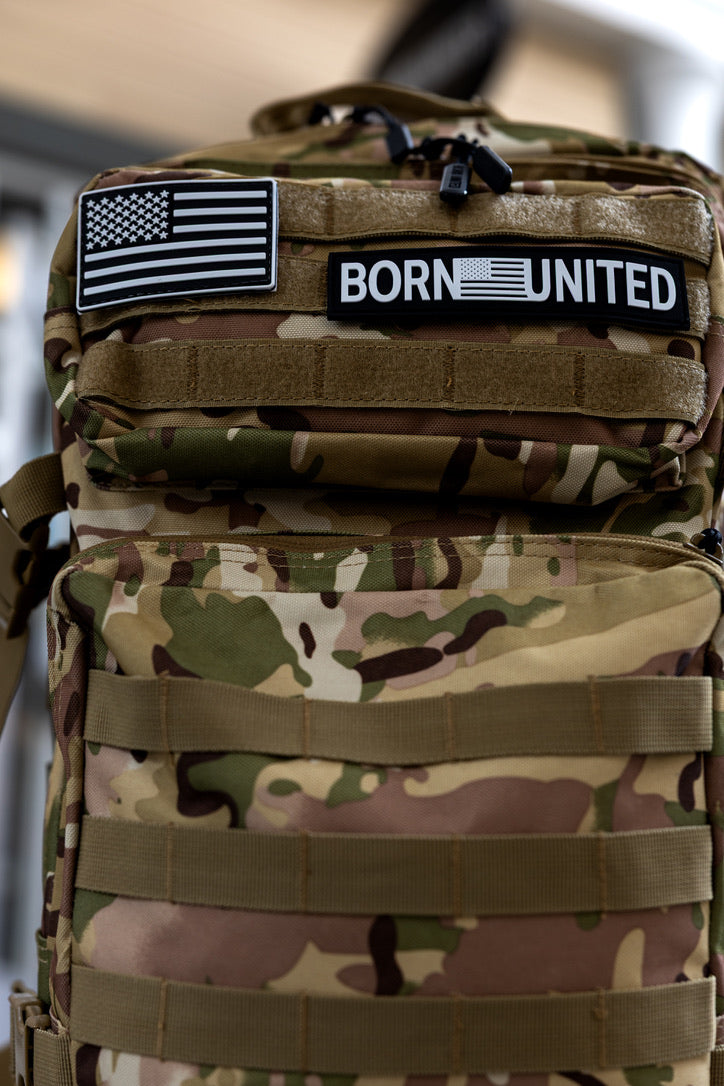 The Born United Private Label OCP Tac-Pac - 45L features a camouflage design, two front patches (black-and-white American flag and BORN UNITED), multiple straps, and a zippered pocket.