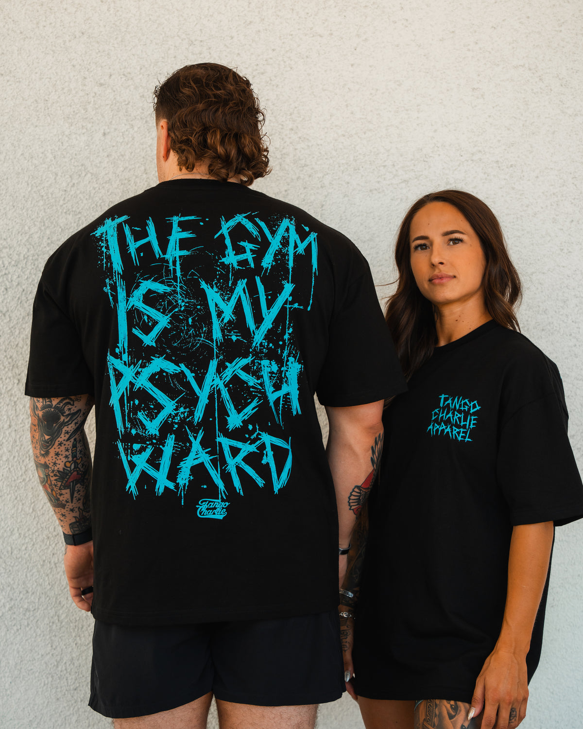 A man and a woman wear Tango Charlie Apparels Psych Ward - Premium Oversized Tee in black; his says THE GYM IS MY PSYCH WARD in bold blue, while hers features the brand logo in smaller blue text on heavyweight cotton.
