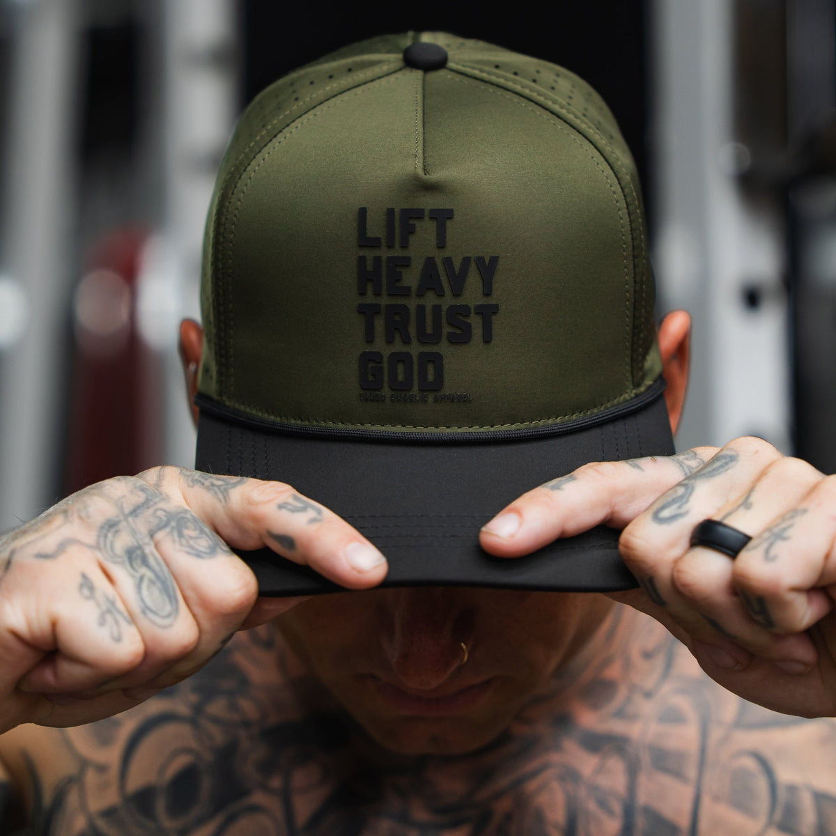 A tattooed person wearing a Tango Charlie Apparel Lift Heavy. Trust God. Hydro - OD Green Snapback Hat while holding the brim and looking downward.