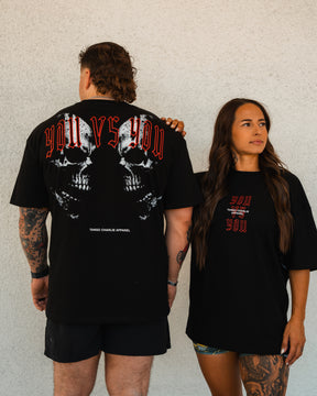 A man and woman wear Tango Charlie Apparel’s You vs You - Premium Oversized Tee. The man displays the skull back graphic, while the woman faces forward, showing the unisex tee’s front design against a plain wall.