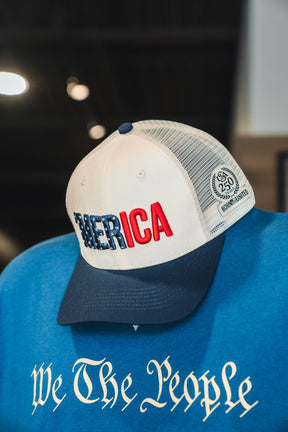The Born United Private Label ’Merica Trucker Snapback—featuring blue and red “MERICA” lettering on a blue-and-white mesh cap—rests atop a blue shirt with “We The People” in classic script. Its breathable design ensures all-day comfort.