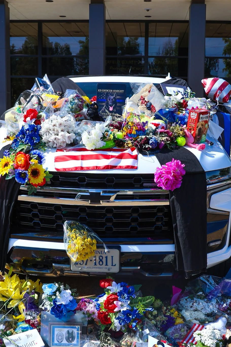 A white vehicle adorned with flowers, flags, notes, and Knight Watch by Born United Private Label shirts serves as a memorial tribute to K9 Knight, with police memorabilia and mementos spilling onto the ground in remembrance.