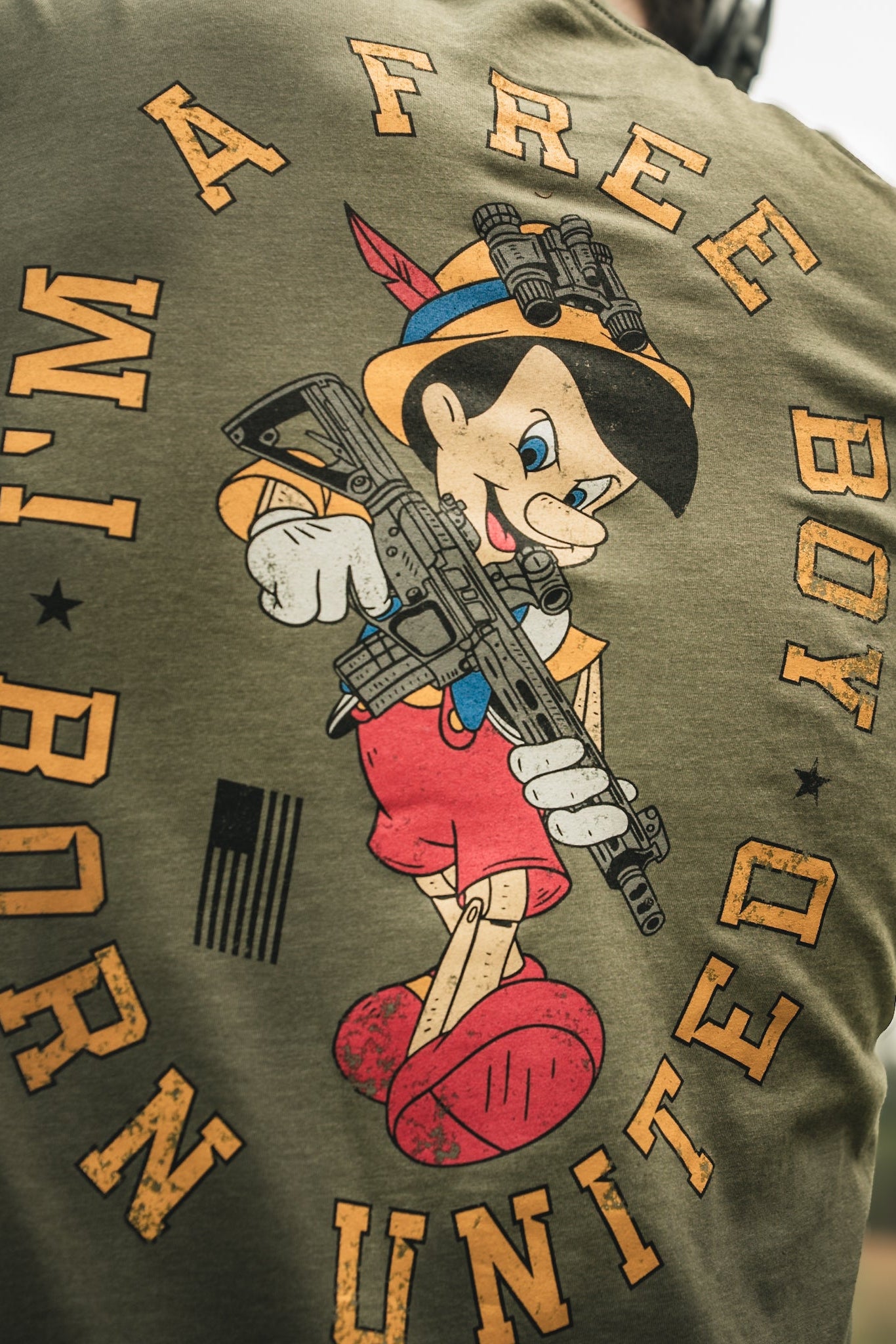 A person wears the Free Boy Tee by Born United Private Label, featuring a wooden puppet character with a rifle, and the text FREE BOY and BORN UNITED, along with an American flag.