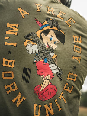 A person wears the Free Boy Tee by Born United Private Label, featuring a wooden puppet character with a rifle, and the text FREE BOY and BORN UNITED, along with an American flag.
