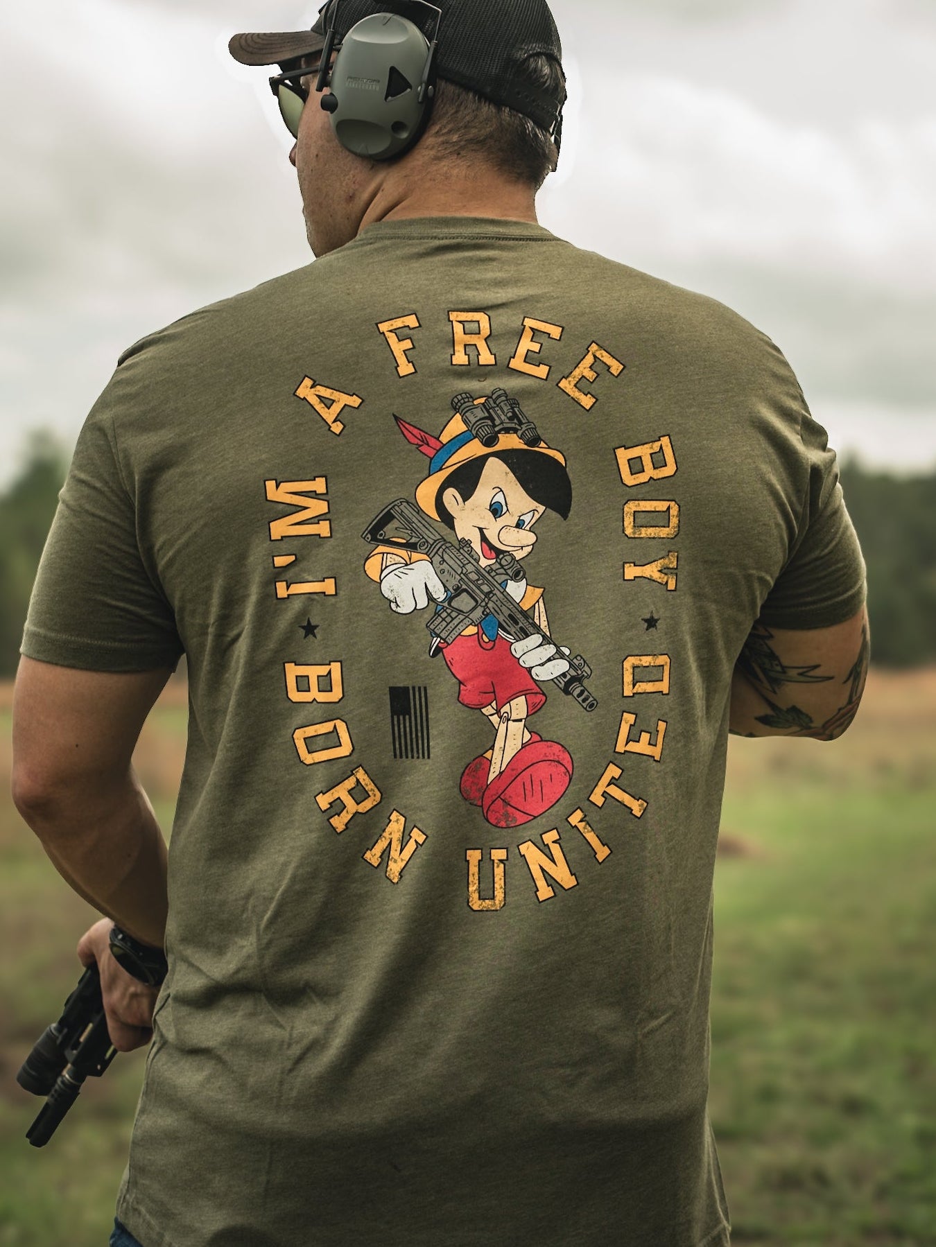 A man outdoors sports headphones and an olive green Free Boy Tee by Born United Private Label, featuring a cartoon character with a gun. The surrounding text reads, Im a free boy but united. Standing in the grass, he confidently holds a gun.