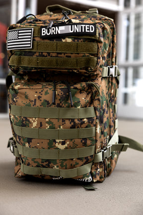 The Digi-Cam Tac-Pac - 45L by Born United Private Label, featuring Marine Corps camo, an American flag patch, and a “BORN UNITED” patch on the front pocket, stands upright on an outdoor surface.