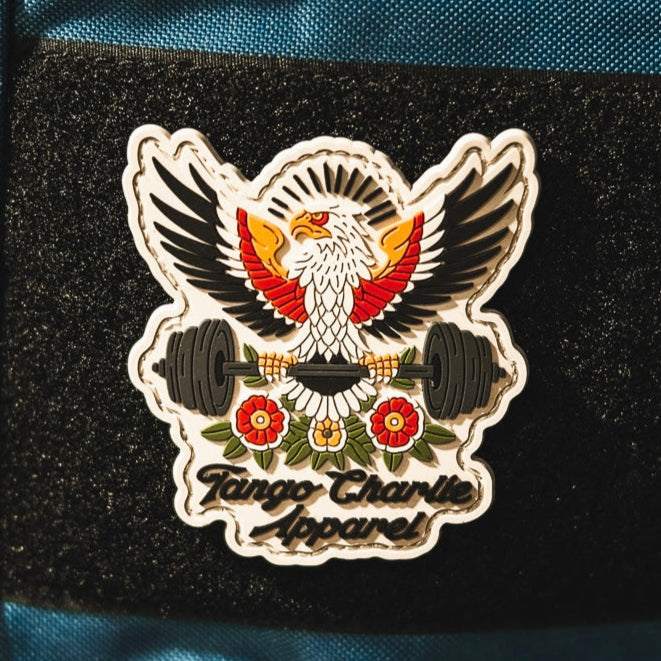 The “Old School, Never Gets Old - Patch” by Tango Charlie Apparel is a 3" 3D PVC patch showing an eagle with outstretched wings and a barbell, surrounded by flowers. The patch displays the brand name and is shown on black fabric.