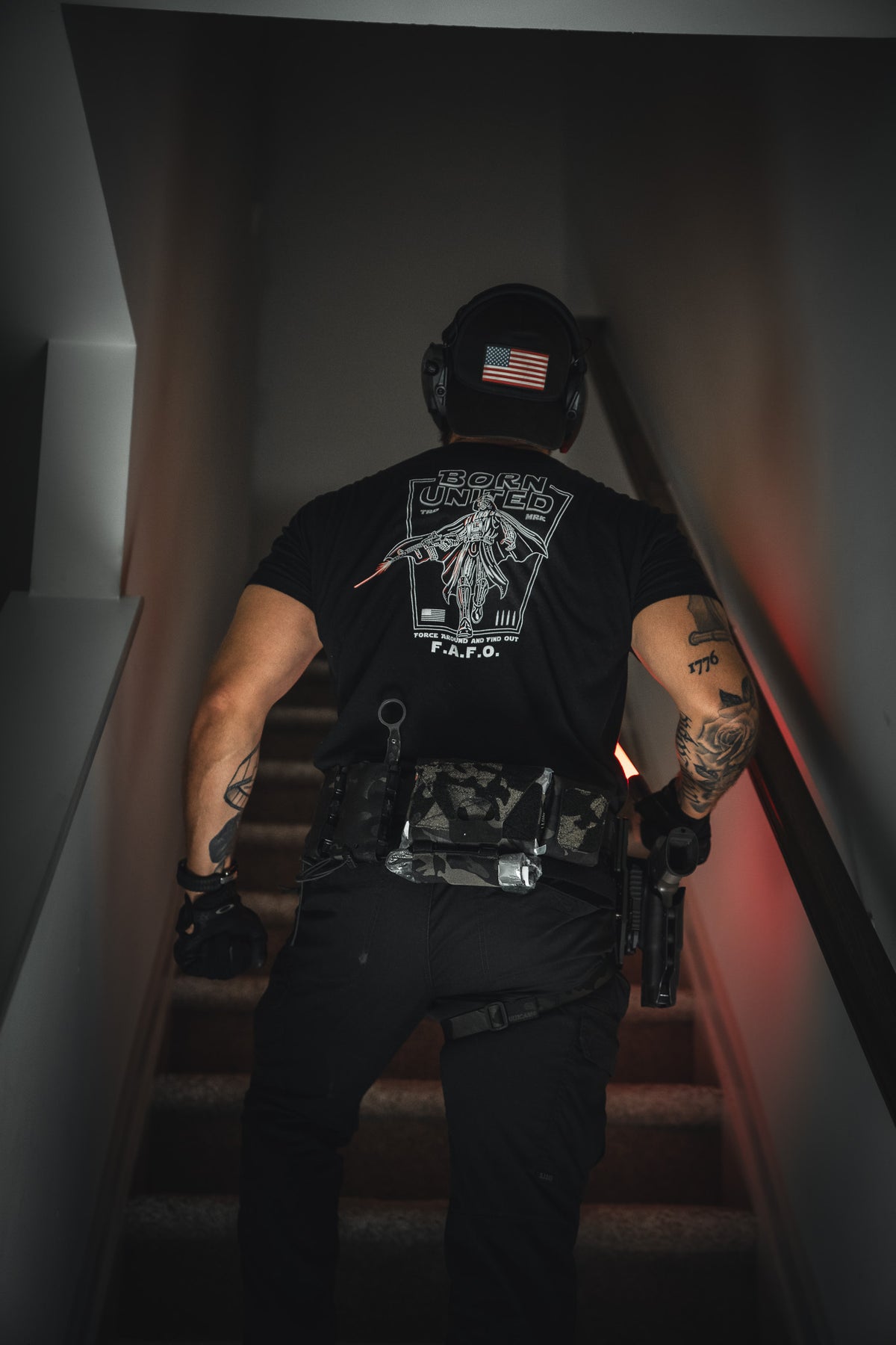 Wearing the Vader FAFO graphic tee by Born United Private Label, a figure in tactical gear with gloves and an American flag helmet patch stands on a stairway, gun holstered—a true Force Around and Find Out attitude.