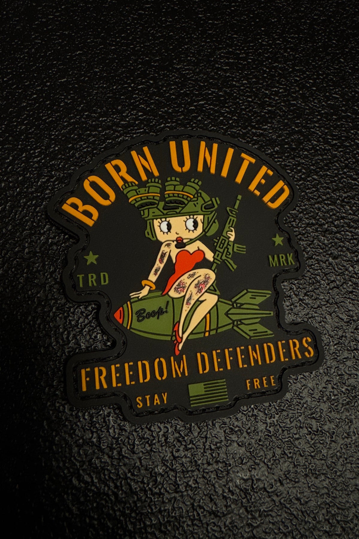 The Born United Private Label Betty Boop PVC Patch features a cartoon pin-up girl in military gear riding a bomb, with “Freedom Defenders,” “BORN UNITED,” “STAY FREE,” and “Boom!” text on a textured black background.