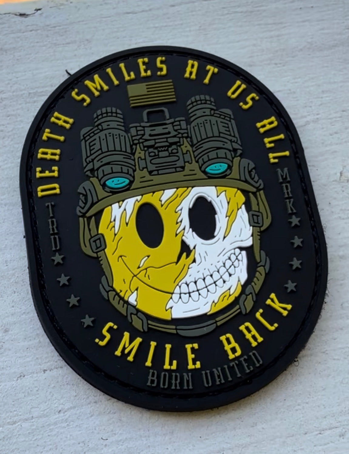 The Smile Back PVC Patch by Born United Private Label shows a helmeted face—half yellow smiley, half skull with night vision goggles—and the words: Death smiles at us all, smile back. Born United—a symbol of stoic resilience.