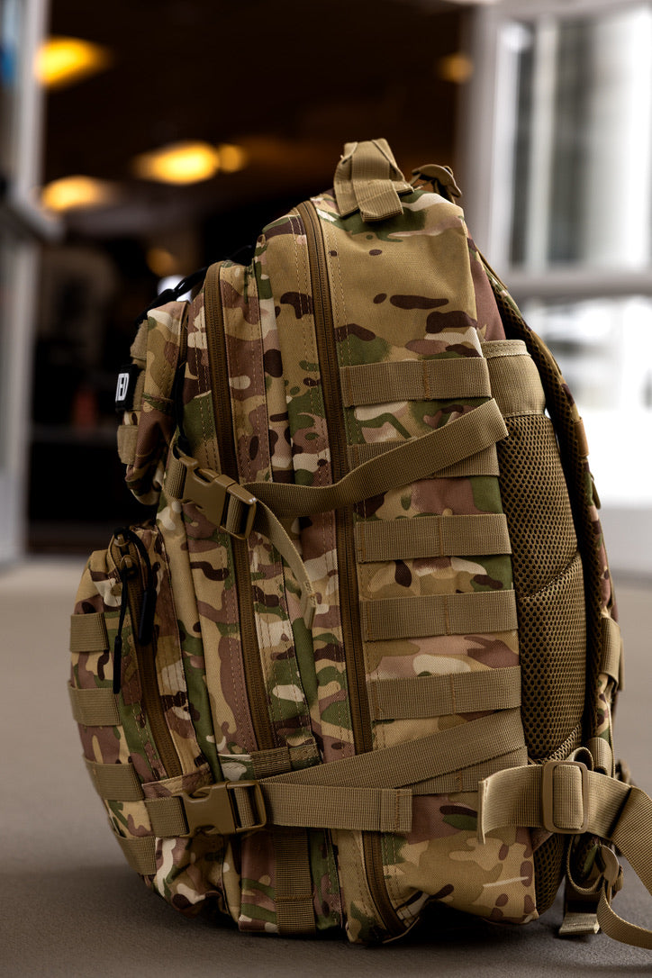 The Born United Private Label OCP Tac-Pac - 45L tactical backpack in tan and green camouflage with multiple compartments and straps stands upright on a smooth indoor floor, with softly blurred open doors and warm lighting in the background.