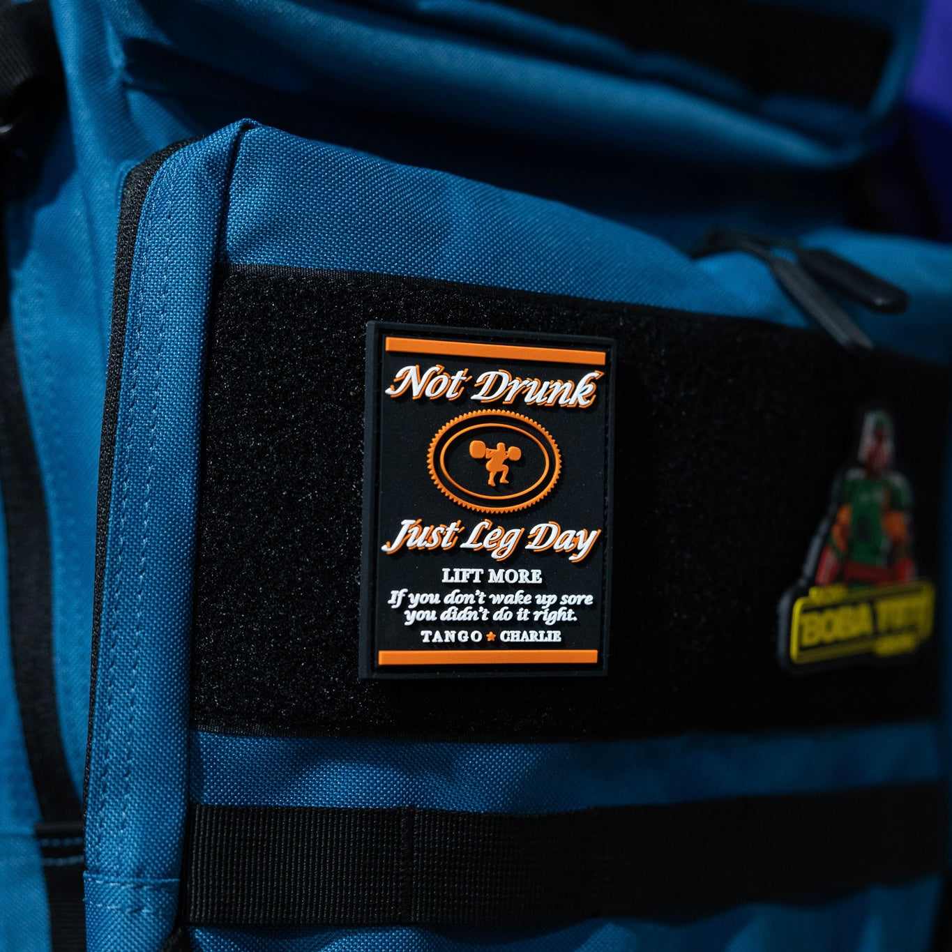 A close-up of the "Not Drunk, Just Leg Day" patch by Tango Charlie Apparel—black with 3D PVC text, LIFT MORE slogan, weightlifter illustration, and Velcro backing.