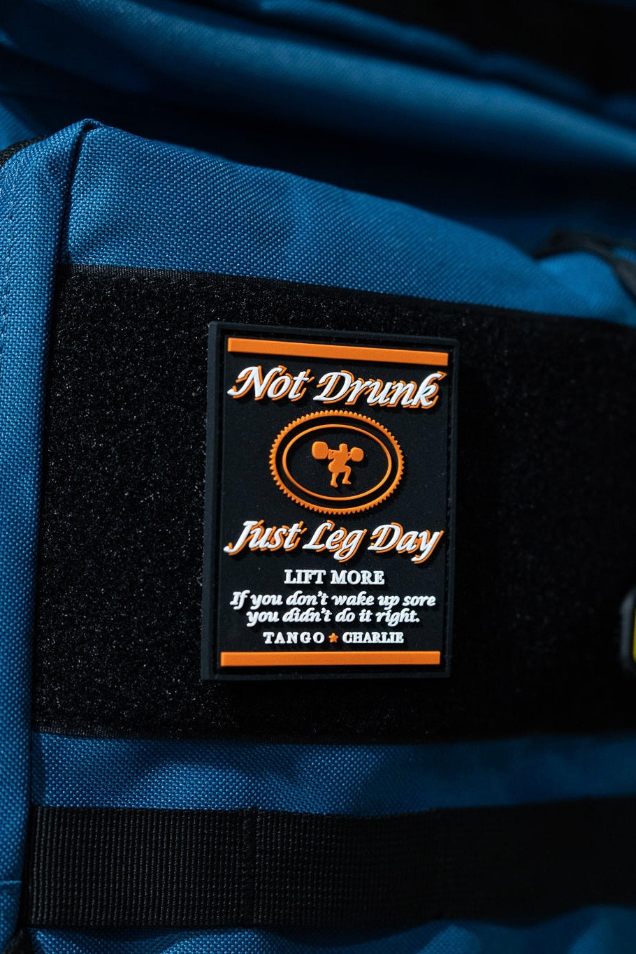 A close-up of the "Not Drunk, Just Leg Day" patch by Tango Charlie Apparel—black with 3D PVC text, LIFT MORE slogan, weightlifter illustration, and Velcro backing.