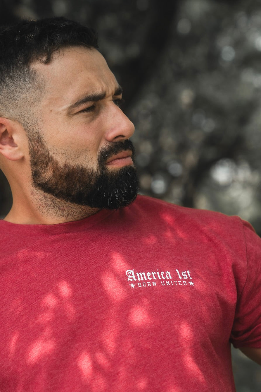 A bearded man with short hair, wearing a red "America First" t-shirt by Born United Private Label, looks to his left while standing outdoors with blurred trees in the background.