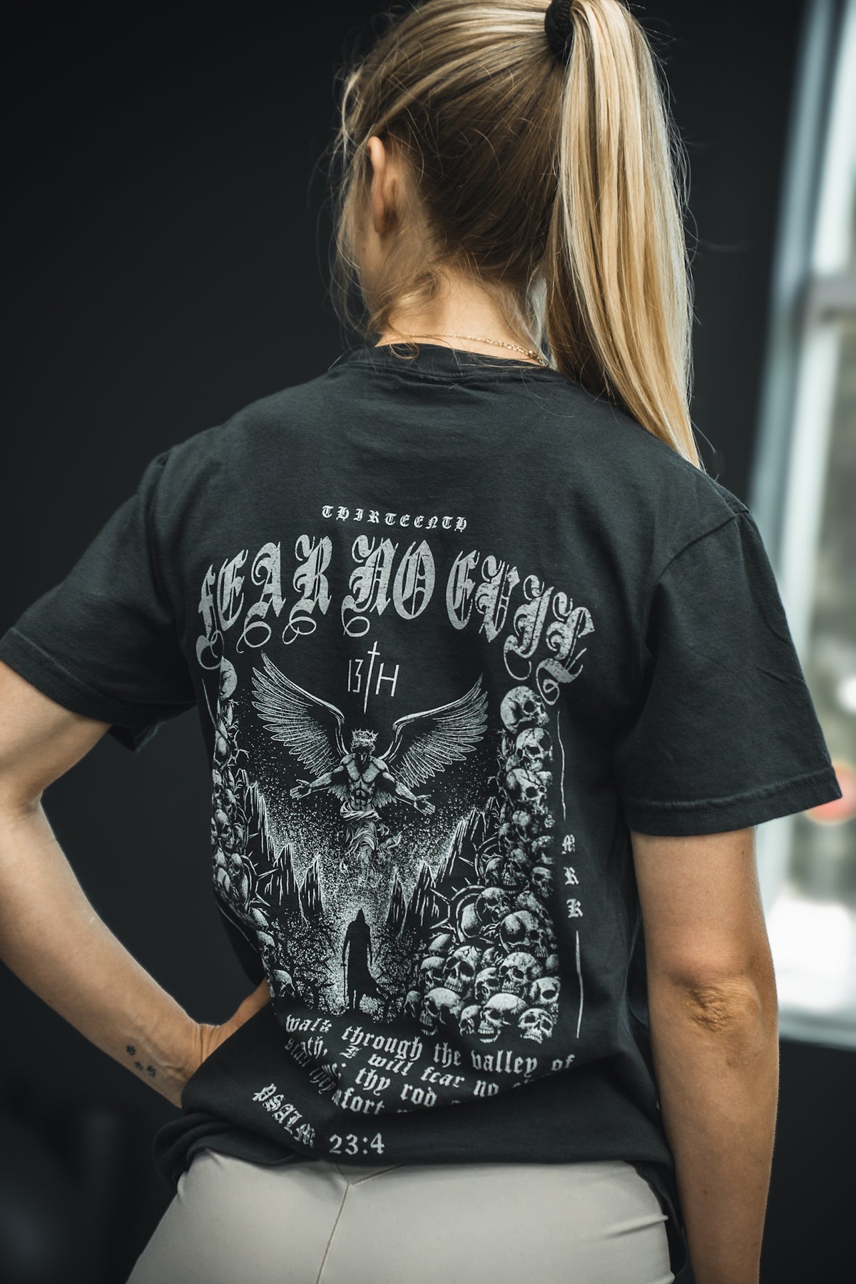 A woman with blonde hair in a ponytail stands with her back to the camera, wearing the 13th Fear No Evil Premium Tee featuring gothic text Fear No Evil, a Psalm 23:4 reference, and an illustrated design on the back.