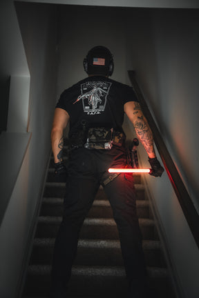 A person in tactical gear sports the Born United Private Label Vader FAFO tee, with tattooed arms, a helmet featuring an American flag patch, and a glowing red baton while standing on a staircase.