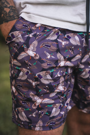 A close-up of Mucker Bucket Men's Performance Athletic Shorts | 5.5"—the Duck Wild shorts in purple with a colorful flying duck pattern. A tattooed arm and white shirt add flair to these versatile athletic gym shorts.