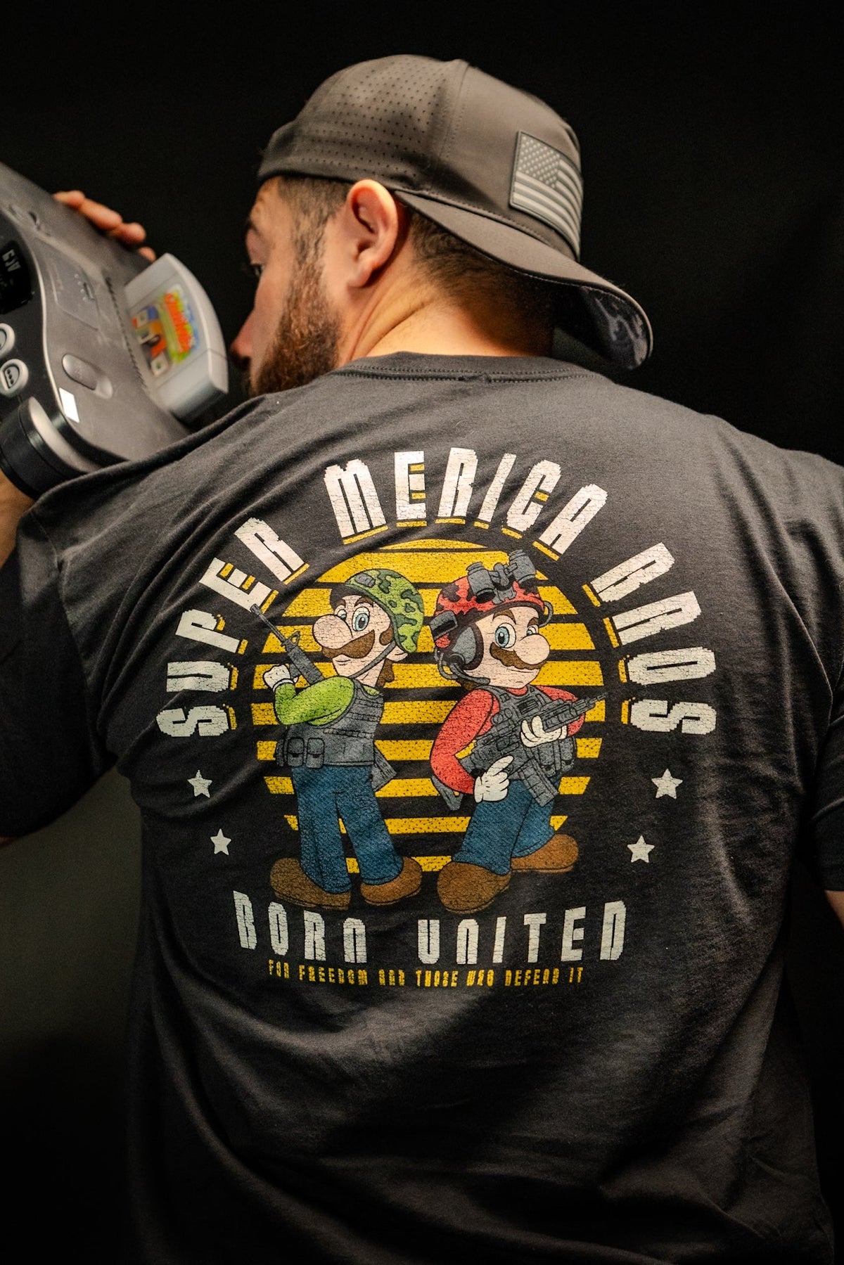 A man in a backwards cap holds an old video game console to his mouth. Hes wearing a premium tee from Born United Private Label, called Super Merica Bros, featuring cartoon plumbers and the slogan For Freedom and Those Who Defend It, celebrating an all-American update of Brothers In Arms.