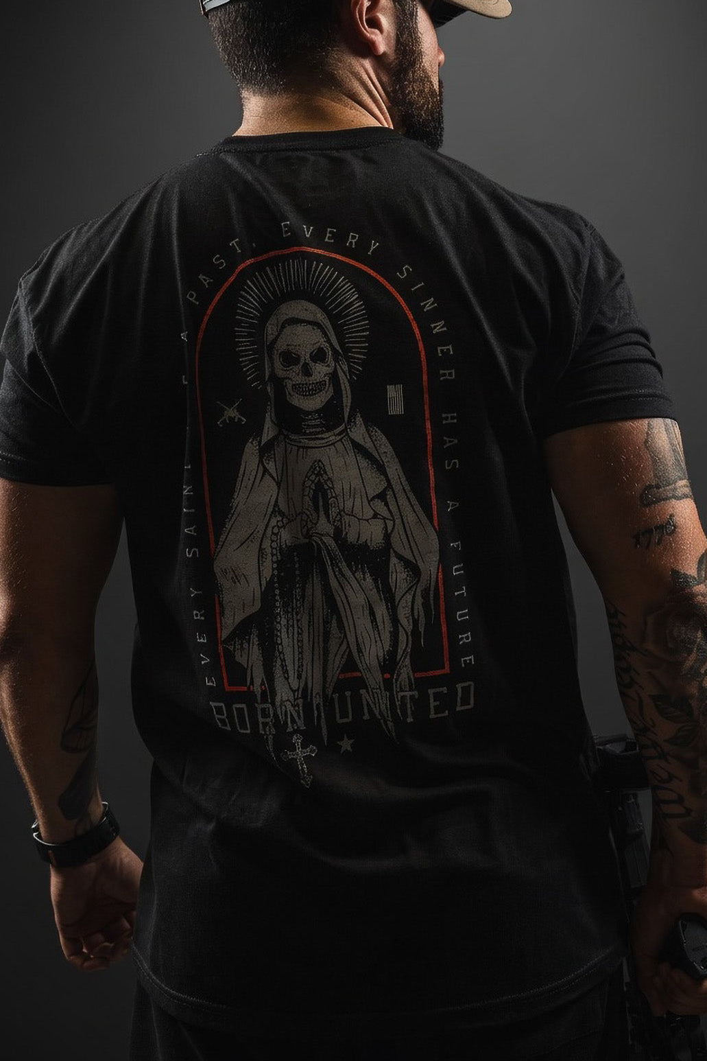 A man wears the Saints and Sinners OG Tee by Born United Private Label—a black shirt with a saintly skeleton and the text: “Every saint has a past. Every sinner has a future. Born United” on the back, plus a matching black cap.