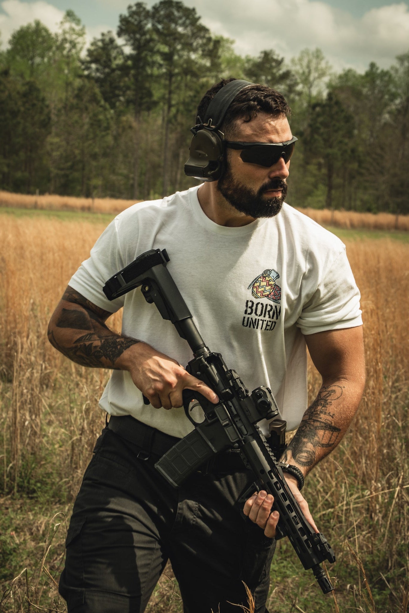 A man in sunglasses, ear protection, and a white Born United Private Label "Battle Bunny" t-shirt stands in a grassy field with a black rifle; trees and clouds form the background.