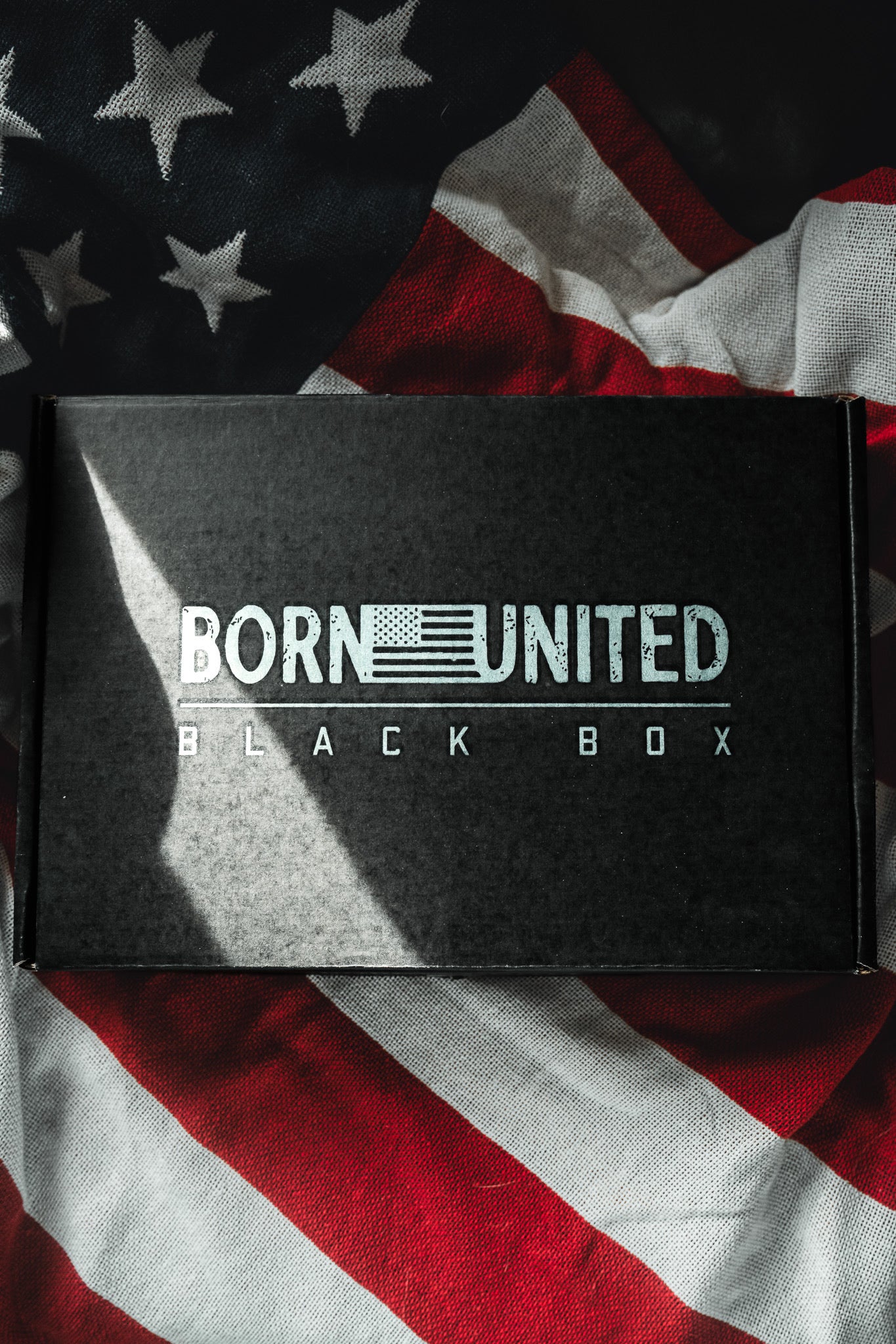 The Born United Private Label "Born United Black Box - Tactigator" sits atop a wrinkled American flag, sunlight highlighting its mysterious appeal and hinting at the excitement of exclusive or limited edition tees inside.