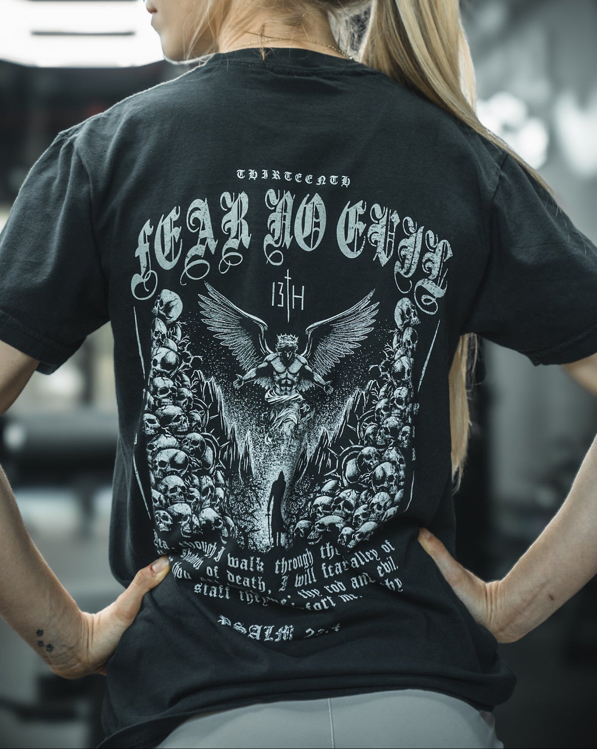 At the gym, a woman displays the back of her black 13th Fear No Evil Premium Tee, featuring an angel, skulls, and “FEAR NO EVIL”—a design inspired by Psalm 23:4 for comfort and strength.