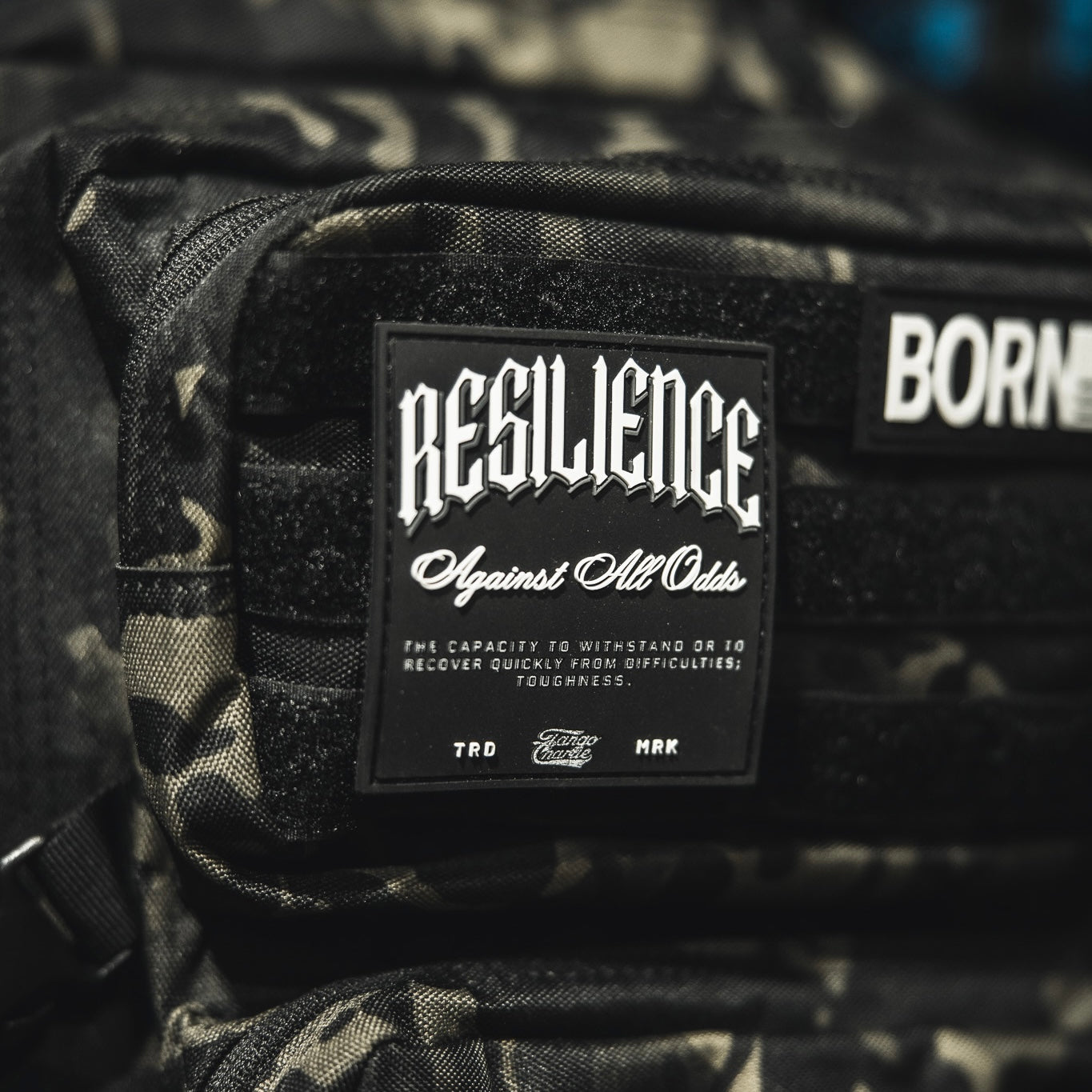 A close-up of the Tango Charlie Apparel "Resilience - Patch," a 3D PVC patch on a camo bag, featuring the phrase "RESILIENCE Against All Odds" to symbolize toughness and overcoming challenges.