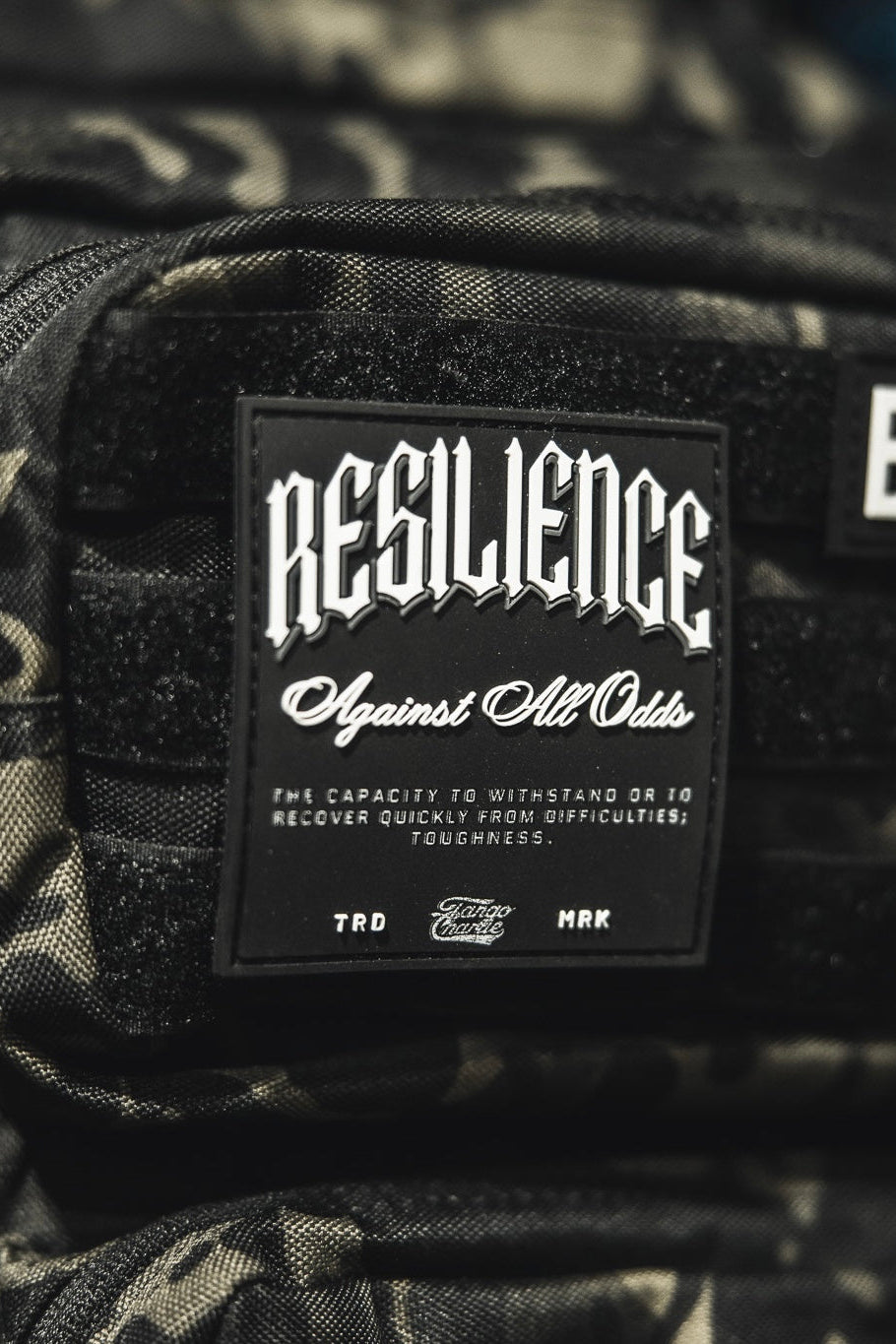 A close-up of the Tango Charlie Apparel "Resilience - Patch," a 3D PVC patch on a camo bag, featuring the phrase "RESILIENCE Against All Odds" to symbolize toughness and overcoming challenges.