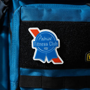 A blue backpack displays a 3-inch ribbon-shaped Patriots Fitness Club patch by Tango Charlie Apparel, in white and blue with a red stripe, attached to a black Velcro section.