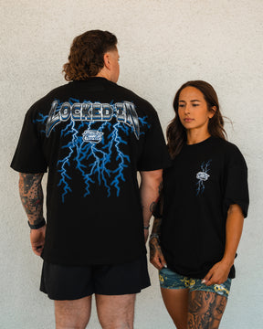 A man shows the back of a Tango Charlie Apparel Locked In - Premium Oversized Tee with blue lightning and LOCKED IN. A woman stands beside him in a comfy unisex tee and patterned shorts, both against a white wall.